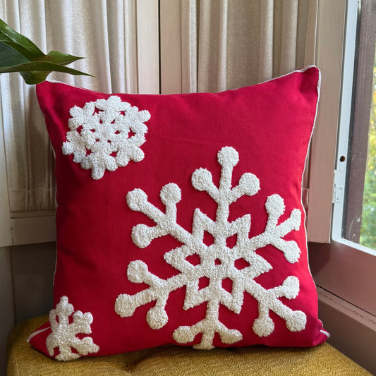 Red pillow cover with large white tufted embroidered snowflakes, styled on a mustard chair near a window with soft natural light and neutral curtains in the background.